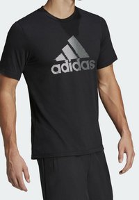 Mustat puuvillapaitat, jossa on heijastava harmaa Adidas-logo, pyöreä kaula-aukko, lyhyet hihat ja rento istuvuus. Sileä rakenne, minimalistinen muotoilu.