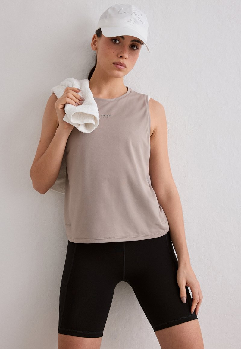 Femme portant un haut beige sans manches, un cuissard de cyclisme noir et une casquette blanche, tenant une serviette blanche sur son épaule contre un mur blanc.