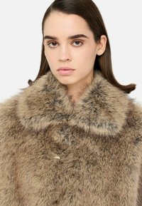 Cappotto in pelliccia sintetica in tonalità marrone chiaro, caratterizzato da un ampio colletto morbido, superficie testurizzata e chiusura a bottone sul collo.