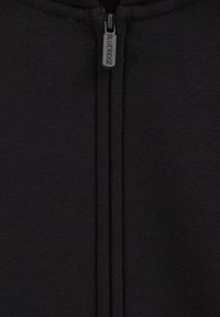 Melnā zipa jaka, izgatavota no mīksta auduma, ar centrālo rāvējslēdzēju un ar zīmola piekabi, kā arī gludu tekstūru.