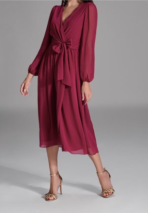 Femme portant une robe midi bordeaux à manches longues avec un nœud à la taille et des sandales à talons hauts dorées, se tenant devant un fond gris uni.