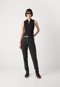 Moteris vilkėti be rankovių juodą zip-up viršų, tamsias džinsus su diržu, bordo spalvos sportinius batus ir juodus saulės akinius, stovinti su viena ranka ant klubų.