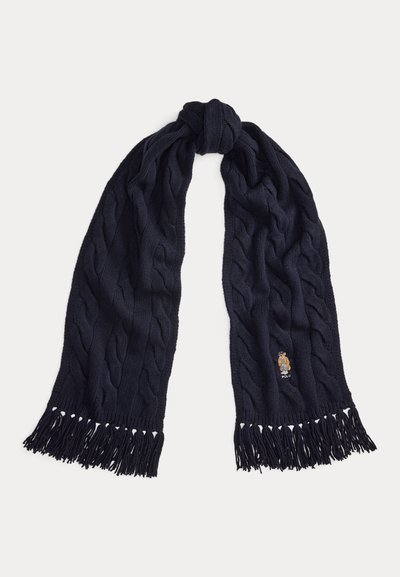 Cachecol azul marinho de malha com padrão de trança, extremidades com franjas e um pequeno logotipo bordado com um urso perto da borda inferior.