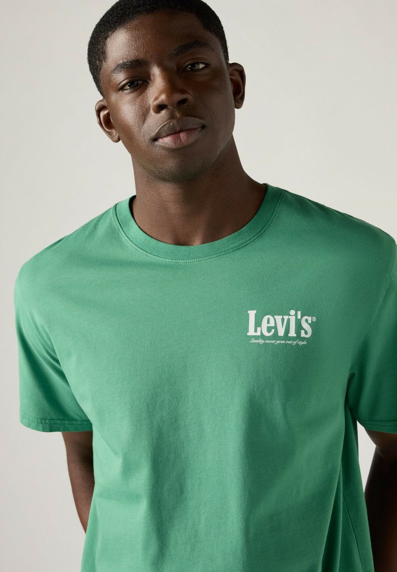 Junger Mann trägt ein grünes Levi's T-Shirt mit weißem Logo-Schriftzug und steht vor einem schlichten hellen Hintergrund.