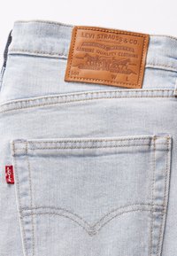 Světle modré džíny z denimu s koženou nášivkou s vyraženým textem, se šitými detaily a červenou tabulkou na zadní kapse.
