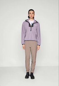Mikina z fleece v svetlo fialovej farbe s čiernymi akcentmi, so štvrťovým zipsom a jednou prednou vreckom, skombinovaná s béžovými nohavicami a čiernymi topánkami.