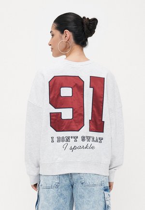 Grå hettegenser med stort rødt "91" på ryggen og hvit tekst som sier "JEG SVEITER IKKE, JEG GLITRER." Har en avslappet passform og ribbede mansjetter.