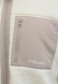 Giacca in pile bianca con una tasca in tessuto beige. Presenta una chiusura con zip e il logo "JJ Studios" in grigio chiaro sulla tasca.