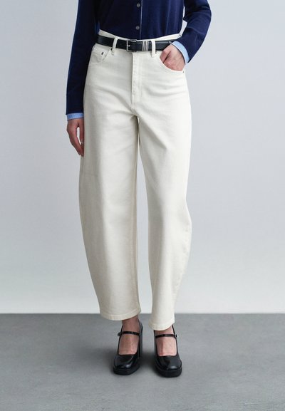 Personne portant un pantalon blanc taille haute à jambes larges avec une ceinture noire, un pull boutonné bleu marine par-dessus une chemise bleu clair, et des chaussures Mary Jane noires.
