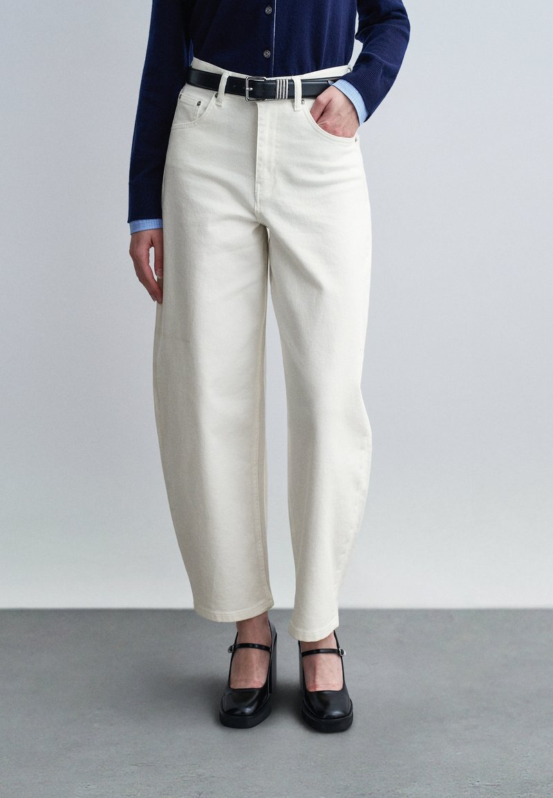 Άτομο που φοράει ψηλόμεσο λευκό παντελόνι με φαρδύ πόδι και μαύρη ζώνη, σκούρο μπλε πουλόβερ με κουμπιά πάνω από ένα ανοιχτό μπλε πουκάμισο, και μαύρα παπούτσια Mary Jane.