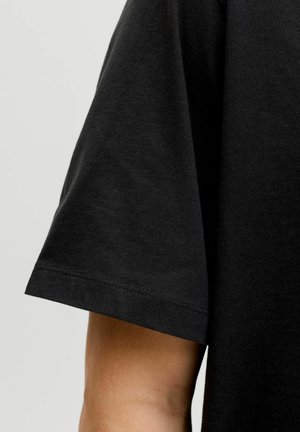 Close-up of a person's arm wearing a plain black short-sleeve t-shirt against a neutral gray background.