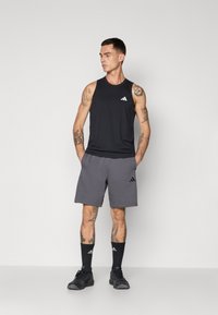 Mand med tatoveringer iført sort ærmeløs atletisk top, grå shorts, sorte sokker og sorte sneakers, stående med hænderne i lommene.