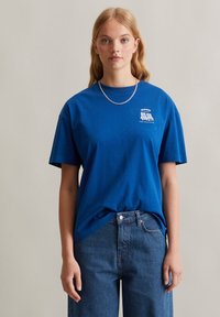 Blaues Baumwoll-T-Shirt mit Rundhalsausschnitt, kurzen Ärmeln und einem kleinen weißen Grafikdesign auf der linken Brust. Kombiniert mit Jeans.