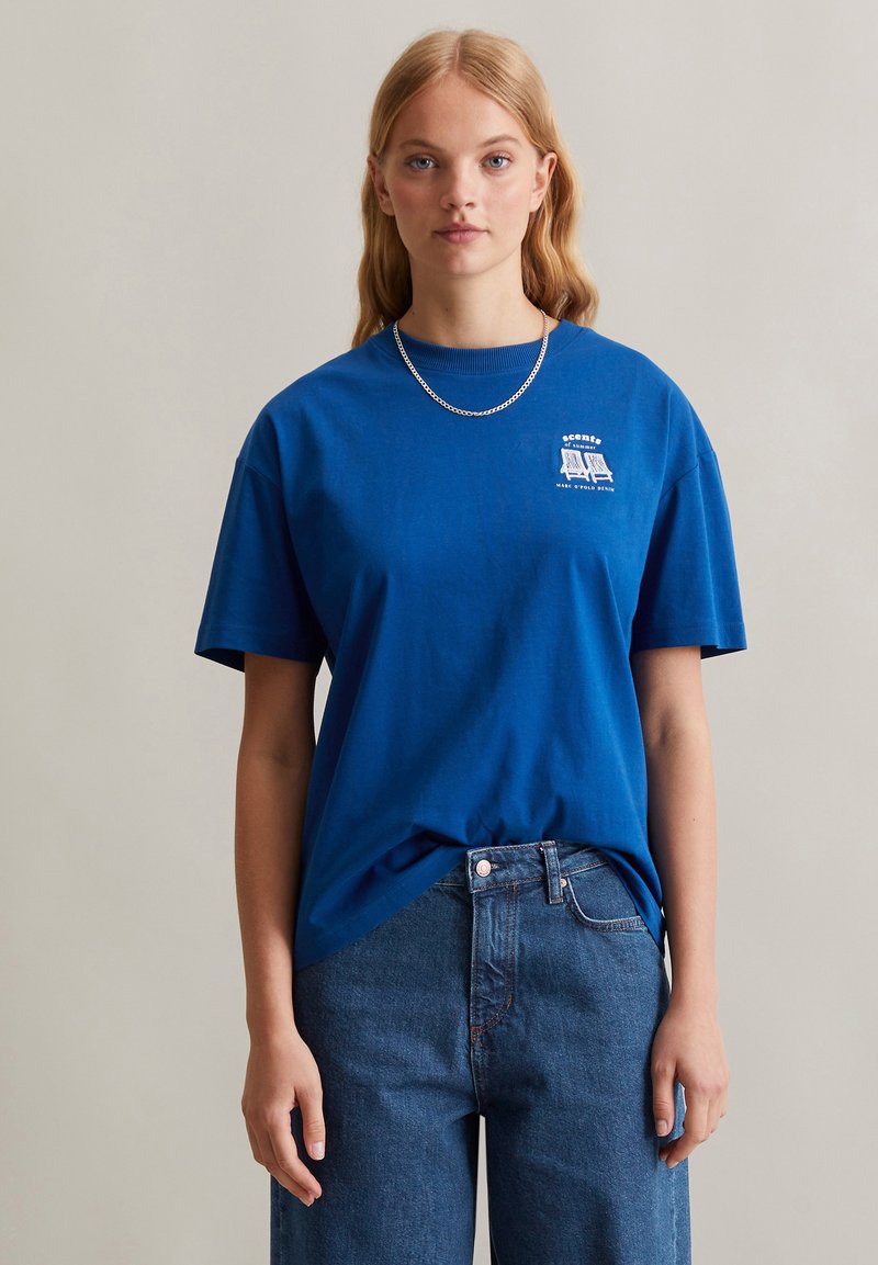 Blaues Baumwoll-T-Shirt mit Rundhalsausschnitt, kurzen Ärmeln und einem kleinen weißen Grafikdesign auf der linken Brust. Kombiniert mit Jeans.