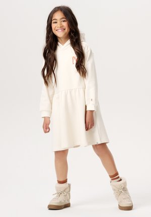 Beige Kapuzenkleid mit geraffter Taille und einem kleinen dekorativen Motiv. Kombiniert mit beigen Stiefeln und hellbraunen gestreiften Socken.