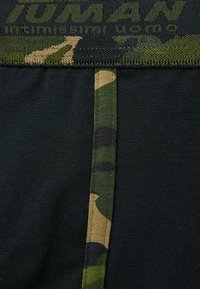 Tela negra con una cinturilla texturizada de camuflaje en verde y beige. Presenta una raya vertical en contraste con diseño de camuflaje.