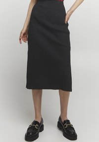 Schwarzer gerippter Midirock mit gerader Silhouette und seitlichen Taschen. Kombiniert mit schwarzen Slippern mit goldenen Akzenten und dicken Sohlen.