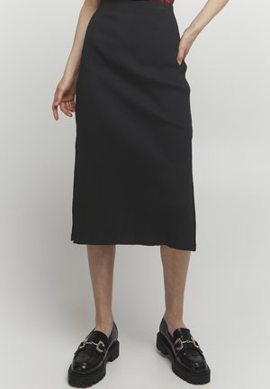 Schwarzer gerippter Midirock mit gerader Silhouette und seitlichen Taschen. Kombiniert mit schwarzen Slippern mit goldenen Akzenten und dicken Sohlen.