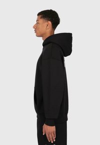 Un jeune homme avec des cheveux bouclés portant un sweat à capuche noir et un pantalon noir assorti, se tenant de profil contre un fond clair.