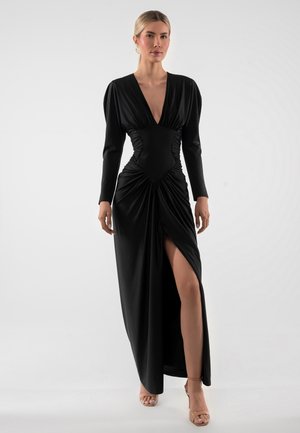 Kobieta stojąca w długiej czarnej sukni z głębokim dekoltem w serek, taliowaną talią, marszczonym dołem i wysokim rozcięciem, ubraną w cieliste szpilki na jednolitym tle.