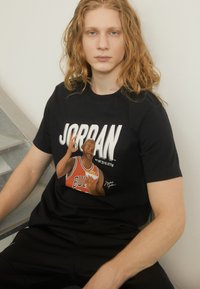 Fekete pamut póló, amelyen egy piros mezben lévő kosárlabdázó grafika látható, a "BULLS" felirattal, valamint nagyméretű fehér "JORDAN" szöveggel a kép fölött.