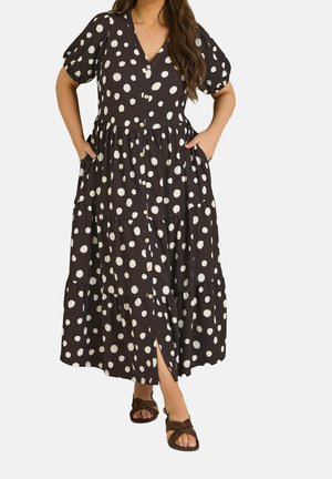 Frau trägt ein schwarzes Midi-Kleid mit weißen Polka-Dots, vorne mit Knöpfen, kurzen Puffärmeln, Seitentaschen und braunen Sandalen.