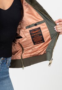 Grüne Bomberjacke mit einem Fellkragen, beigem Satinfutter und einer Innentasche. Das Etikett zeigt "BOMBERS" in schwarzem und orangefarbenem Text.