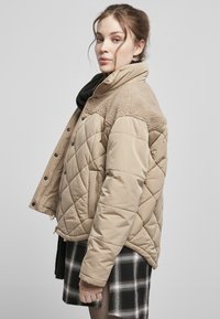 Beige gepolsterte Jacke mit einem gesteppten Design, ausgestattet mit einem Kragen aus Kunstfell und einem Reißverschluss. Getragen über einem schwarz-weißen karierten Rock.