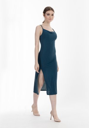 Frau in einem ärmellosen dunkelblauen Midikleid mit Oberschenkelschlitz, die läuft, trägt beige High Heels, vor einem schlichten weißen Hintergrund.