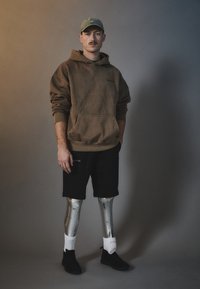 Sudadera marrón de gran tamaño con bolsillo, pantalones cortos negros, piernas protésicas plateadas, calcetines blancos y zapatillas negras; textura suave y colores apagados.
