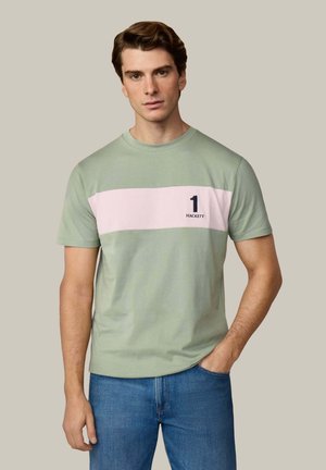 Man draagt lichtgroen t-shirt met een horizontale lichtroze streep en de tekst "1 Hackett", gecombineerd met een blauwe spijkerbroek.