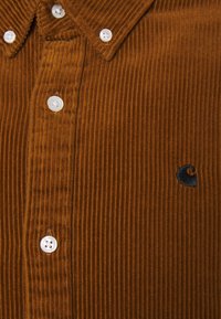 Camisa de pana marrón con estrías verticales, seis botones blancos y un pequeño logo oscuro en el lado izquierdo, cerca de los botones.