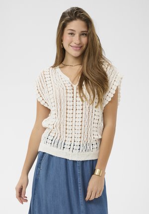 Pikajuukseline noor naine naeratab, kandes valget heegeldatud topi, sinist seelikut, kuldset käevõru ja väikseid kõrvarõngaid, taust on ühtlane.