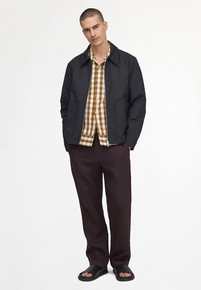 Man wearing black zip-up jacket, beige and white checkered shirt, dark brown pants, and black open-toe sandals, standing against white background.