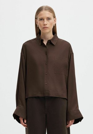 Un mannequin porte une chemise courte marron foncé, boutonnée, avec une poche poitrine et des manches larges, assortie à un pantalon, sur un fond uni.