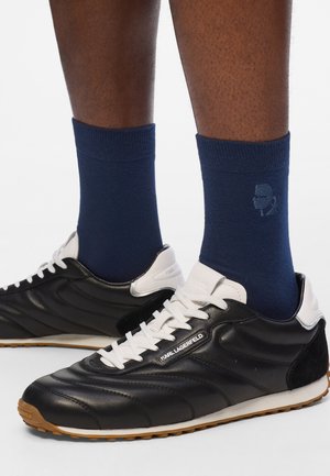 Baskets en cuir noir avec des accents blancs et une semelle en caoutchouc, associées à des chaussettes bleu marine arborant un logo subtil. Texture lisse et design athlétique.