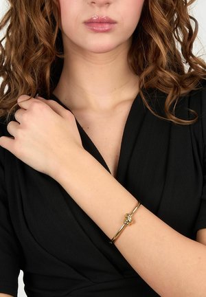 Donna con capelli ricci che indossa un top nero e un braccialetto d'oro con dettaglio a nodo al polso, tenendo la spalla opposta.