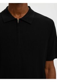 Polo noir à manches courtes avec col et courte fermeture éclair à l'avant, porté par une personne à la peau foncée.