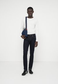 Calças de ganga azul-escuro combinadas com uma camisa branca de mangas longas. Uma mala cruzada azul-escura pende do ombro do modelo. Ténis pretos completam o visual.