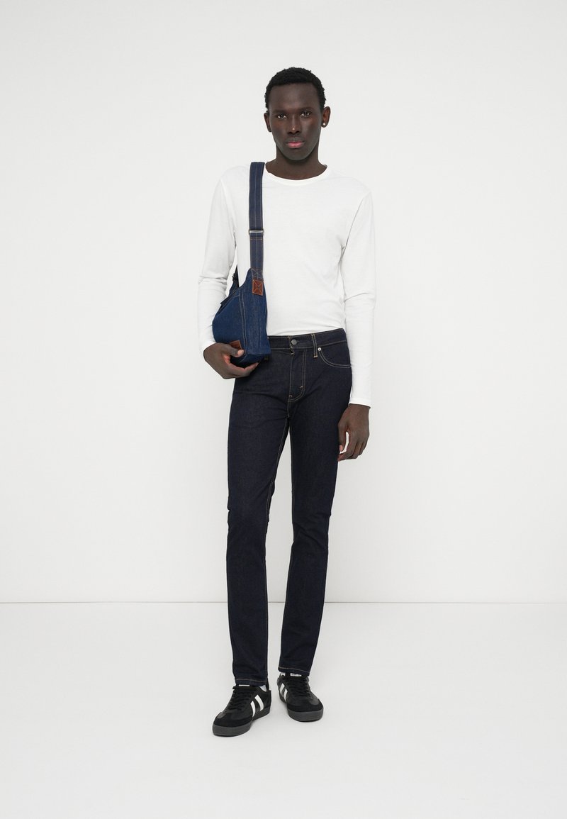 Un jean en denim bleu foncé associé à une chemise blanche à manches longues. Un sac bandoulière bleu foncé pend à l'épaule du modèle. Des baskets noires complètent la tenue.