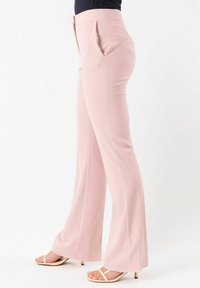 Donna che indossa pantaloni su misura rosa chiaro e sandali beige con tacco, in piedi davanti a uno sfondo bianco.