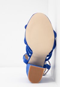 Vue de la semelle d'une sandale bleue en daim à lanières avec un talon bloc, montrant une semelle beige lisse et un dessous de talon texturé.
