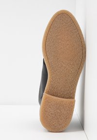 Semelle d'une chaussure noire avec une semelle en crêpe en caoutchouc texturée de couleur tan, appuyée contre un mur d'angle blanc et une surface de sol blanche.