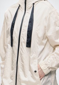 Veste légère couleur crème avec fermeture éclair noire et cordons, poignets élastiques et une main dans la poche du côté droit.