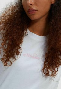 Camiseta blanca de manga corta con un logo rosa "ellesse" en el pecho. La tela parece suave, con una textura lisa y un corte relajado.