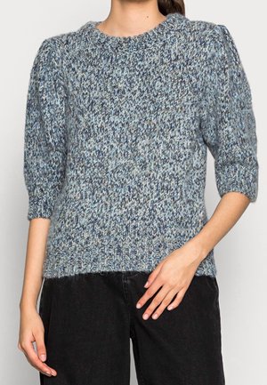 Femme portant un pull en maille bleu et gris avec des manches aux coudes et un pantalon noir, debout avec une main posée sur la hanche.