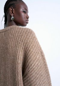 Femme aux cheveux tressés portant un grand pull en maille beige et des boucles d'oreilles argentées avec des pierres vertes, regardant sur le côté sur un fond blanc uni.