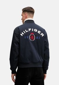 Chaqueta bomber marina con cuello y puños de canalé; presenta la inscripción "HILFIGER" en blanco, una corona roja y un logo de león, con "1985" en azul debajo.