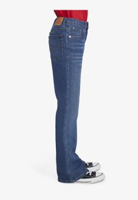 Jean en denim bleu avec une coupe droite, présentant une étiquette en cuir marron, des poches arrière et assortis à des baskets noires.