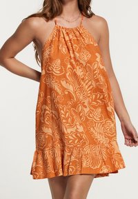Woman wearing sleeveless orange halter dress with light floral print and ruffled hem against white background.